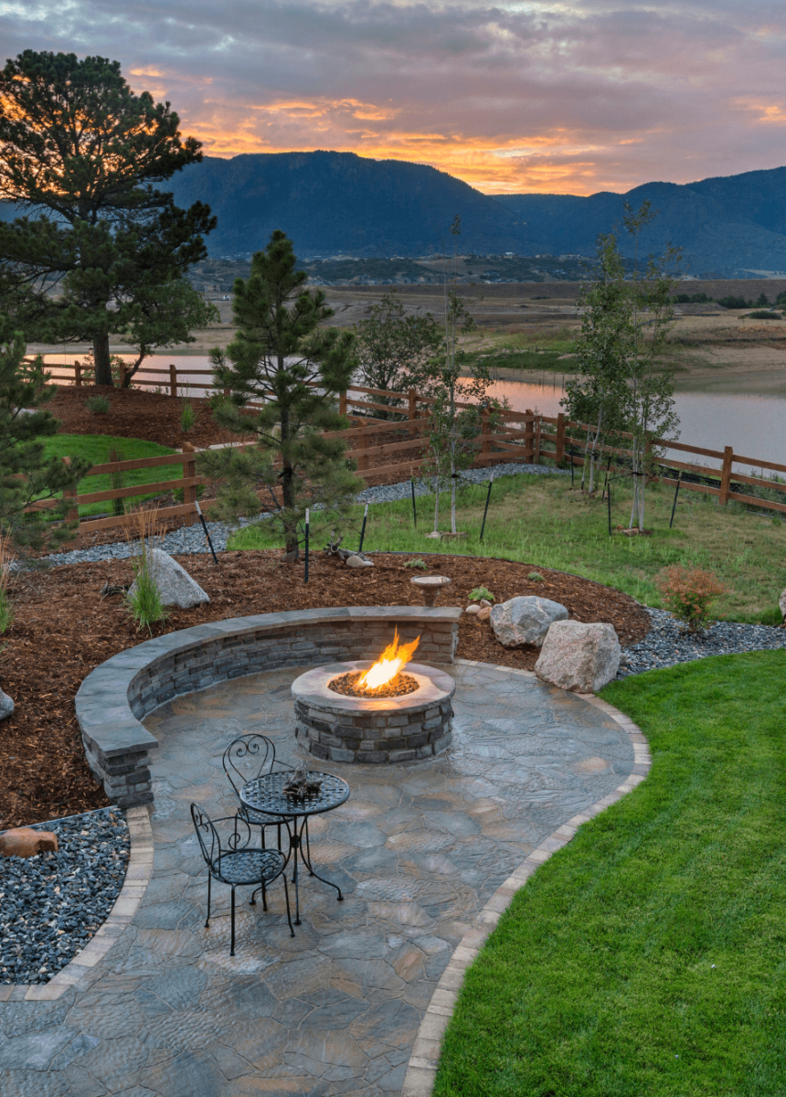 A beautifully landscaped backyard featuring a circular fire pit surrounded by stone patio and seating
