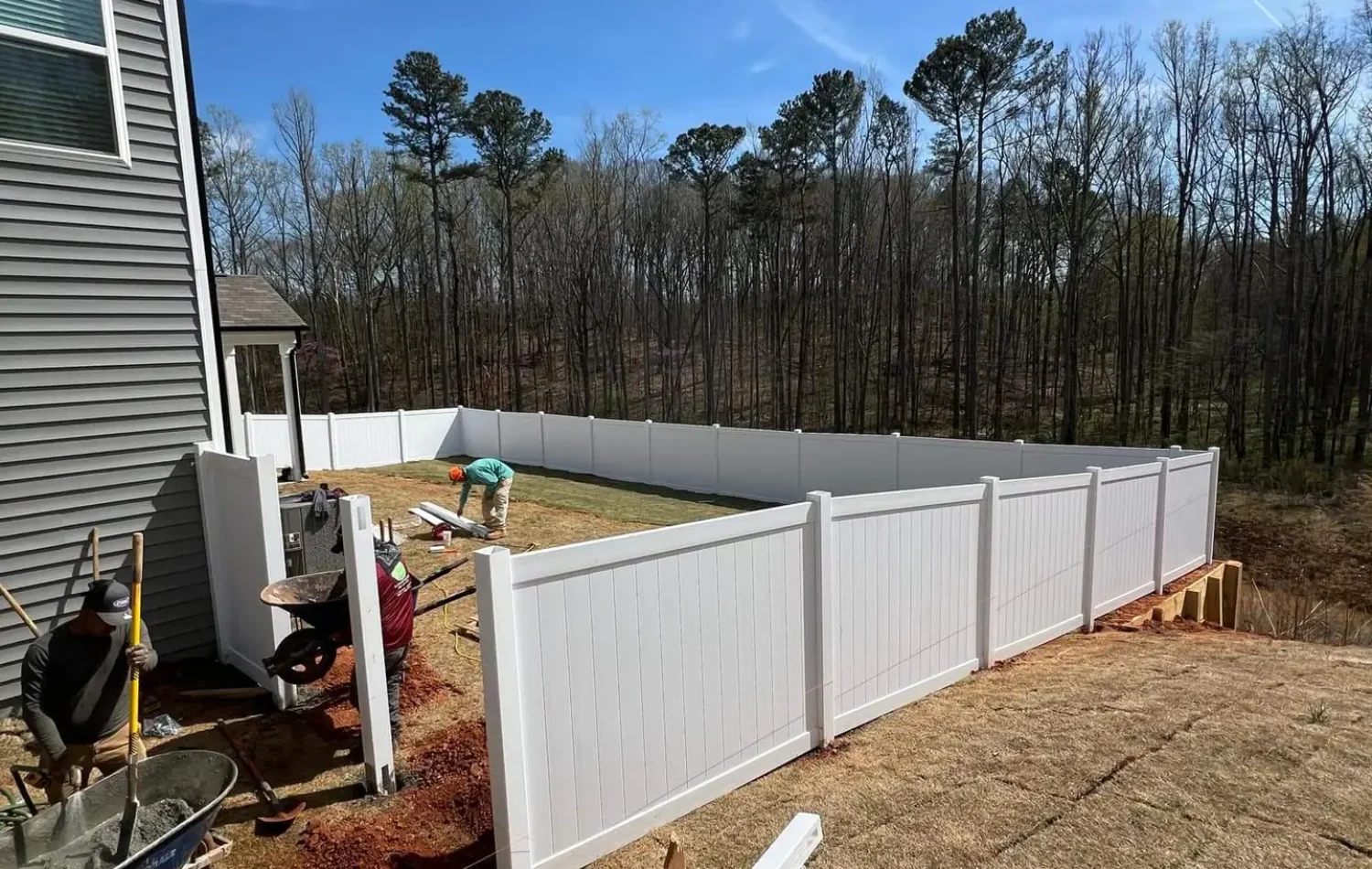 Fence Installation Workers are installing a white vinyl privacy fence in the backyard of a modern gray house.